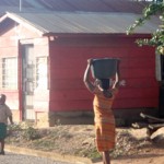 Woman carrying basin on head near red house